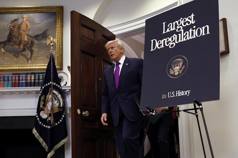 Donald Trump walks into a room wearing a suit next to a sign that says 'Largest Deregulation'.