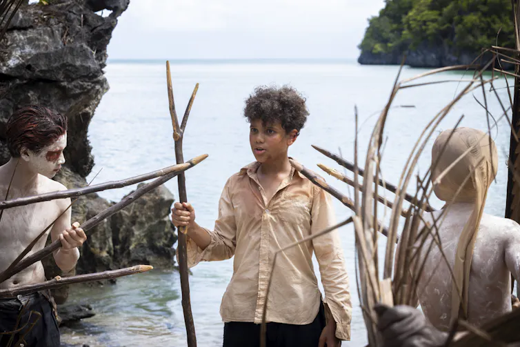 a boy in a shirt, holding a stick spear, surrounded by boys with paint and spears