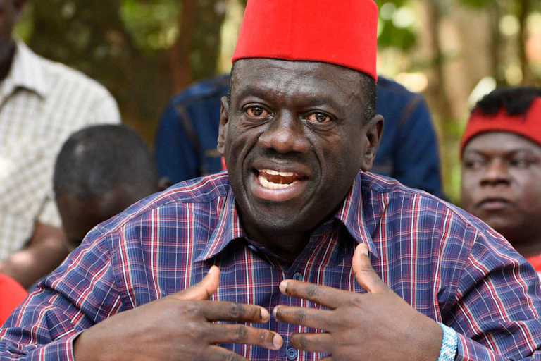 A man dressed in a checked shirt and wearing a red cap on his head gestures at his chest while speaking