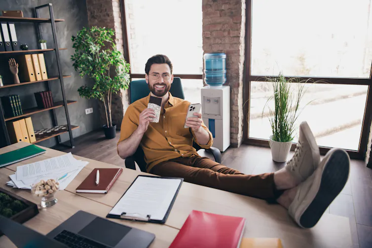A young bearded man in a yellow shirt grins as he holds up a chocolate bar and sits with his feet on an office table.