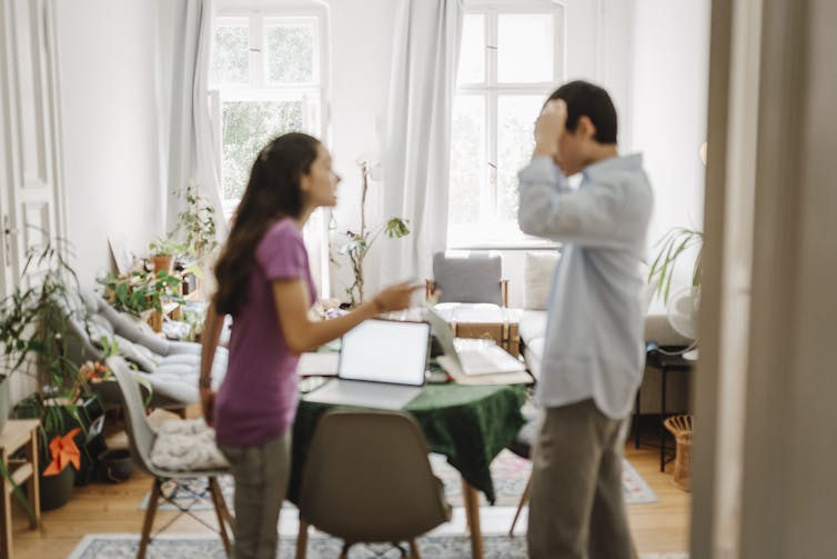 A dark-haired man and woman stand as they seem to argue in a dining room, with the man clutching his temples.