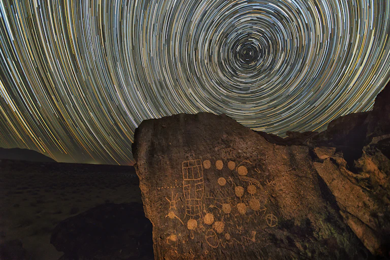 Petroglyphs carved into a rock, with a long exposure of stars circling one point in the background.