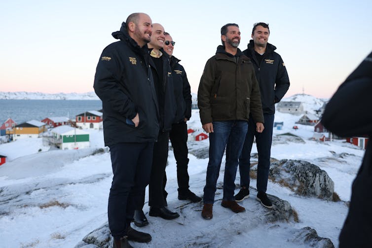 Five white men standing on snow smile for the cameras with a Greenland village behind them.