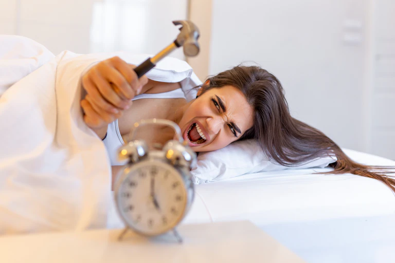 Woman lying in bed about to hit the 5am alarm with a hammer