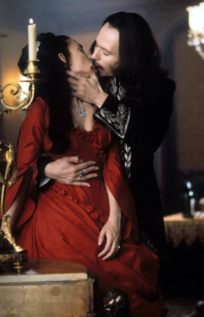 A woman with black hair and a red dress passionately kisses a man with long black hair.