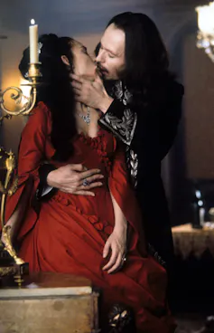 A woman with black hair and a red dress passionately kisses a man with long black hair.