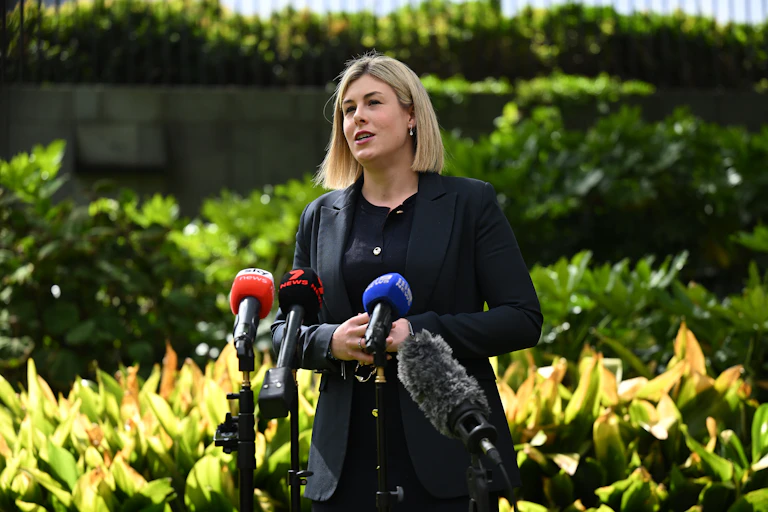A woman with a blonde bob stands in front of a green garden and addresses the media