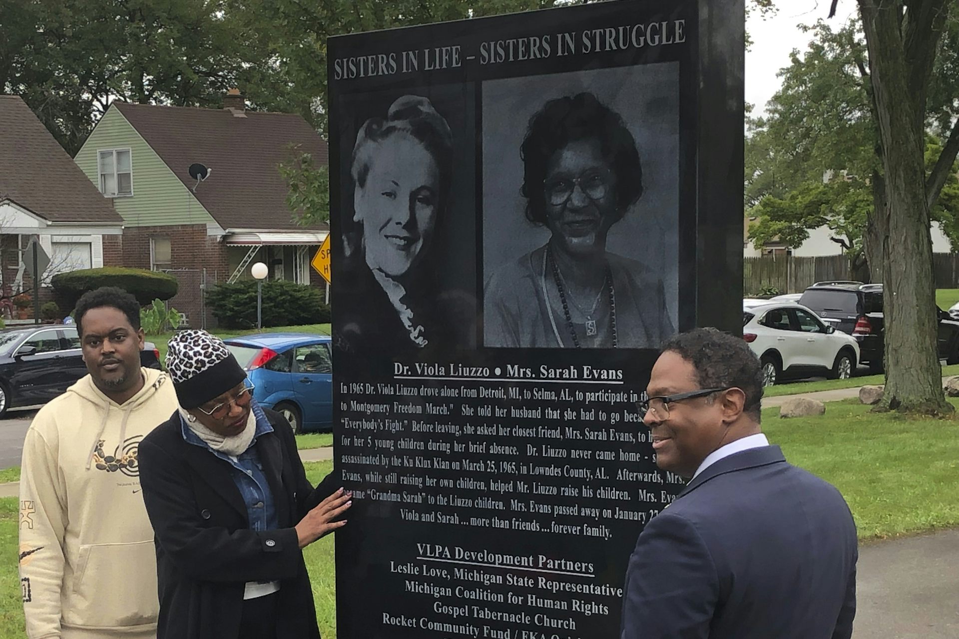 Three people pose for pictures on either side of a black, granite memorial.