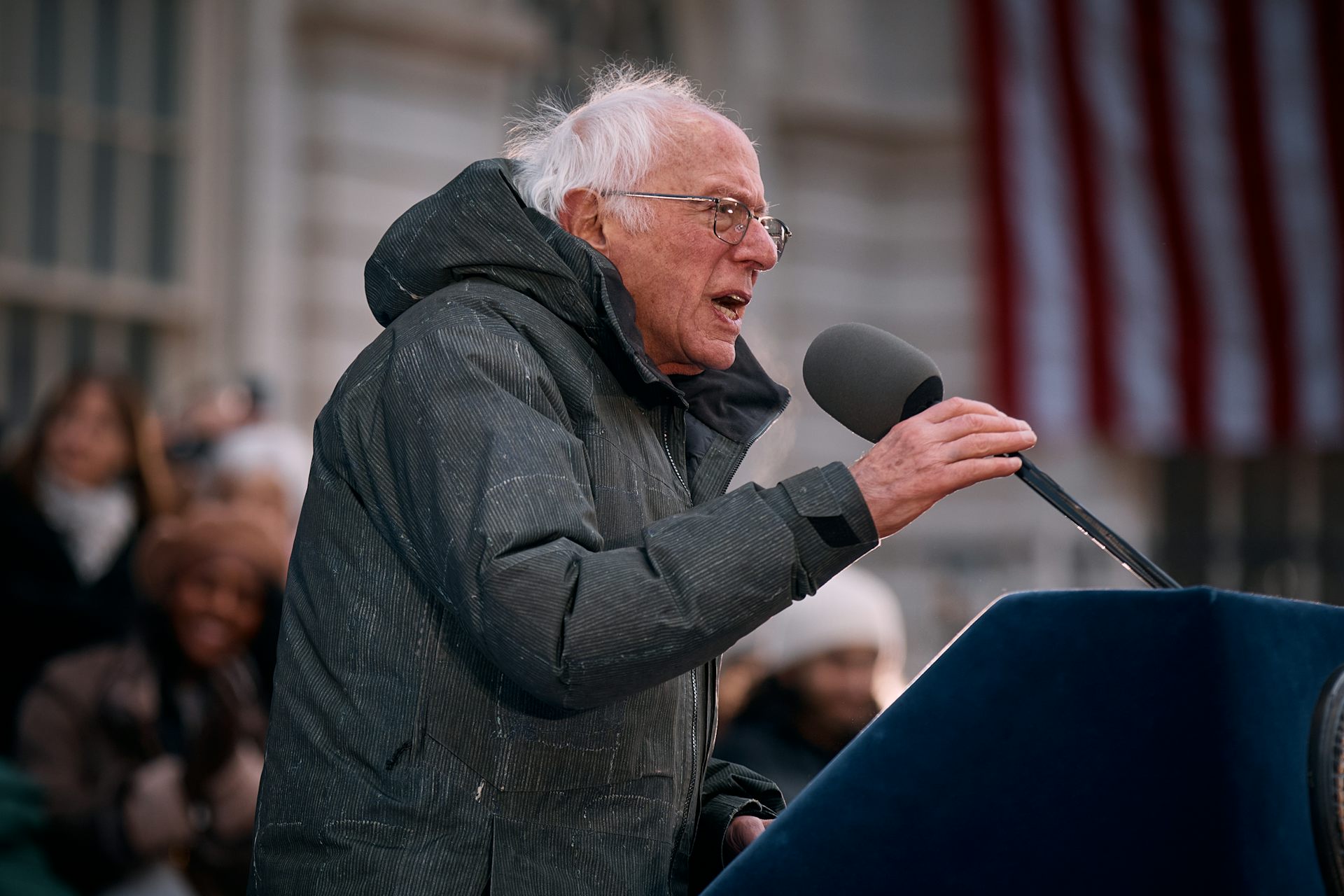 Sen. Bernie Sanders holds and leans into a microphone, wearing a heavy coat at an outdoor event.
