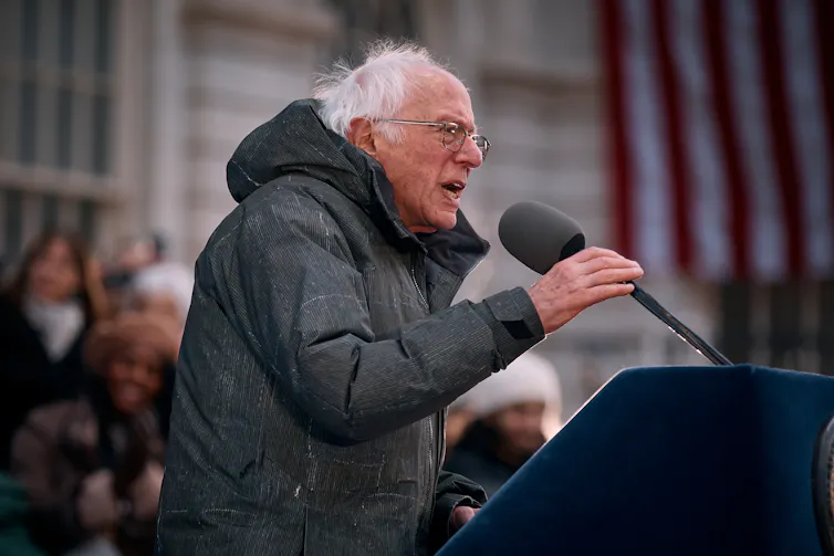 Sen. Bernie Sanders holds and leans into a microphone, wearing a heavy coat at an outdoor event.