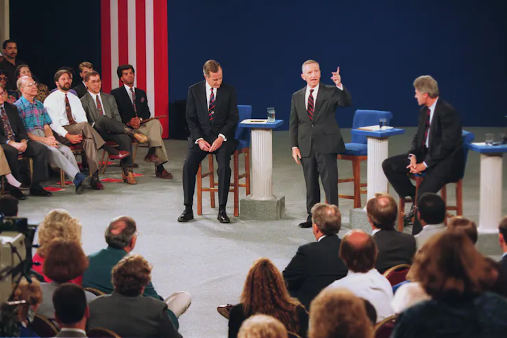 Ross Perot gestures with his left hand while standing between George H.W. Bush and Bill Clinton, both seated on a stage.