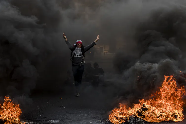 A protestors makes the peace sign as they walk through black smoke, surrounded by burning tires.