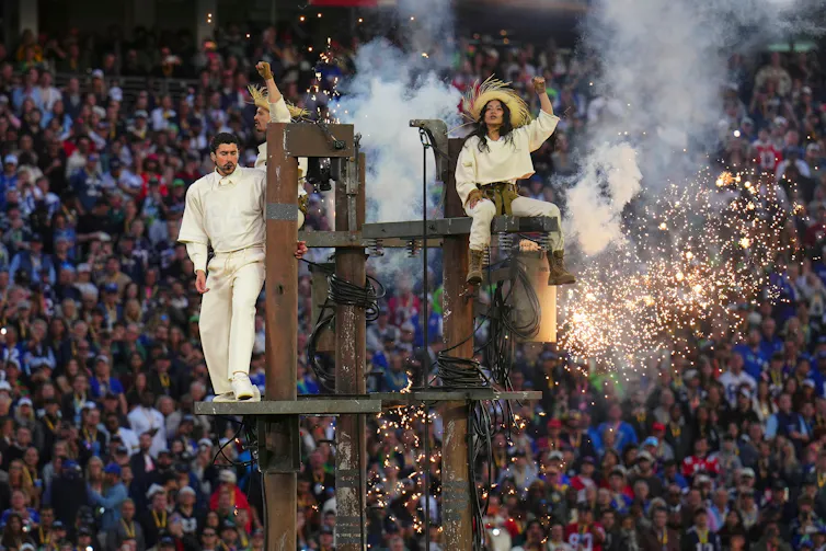 Two people wearing white stand on power poles, with fireworks and a crowd behind them.