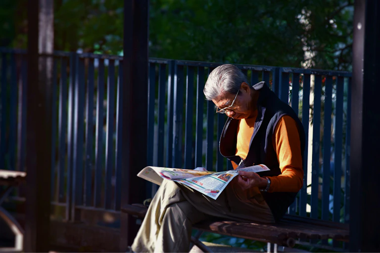 Older man on a bench reading a paper in the sun.