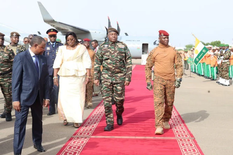 Deux hommes en uniforme militaire marchent sur un tapis rouge avec un avion en arrière-plan.