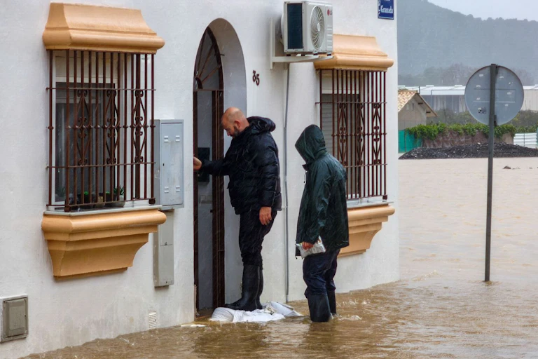 Un hombre abriendo la puerta exterior de una casa, mientras otro que le sigue, en un terreno inundado de agua marrón