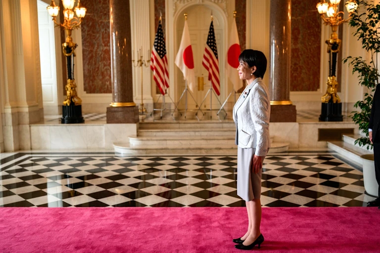 Sanae Takaichi standing on a red carpet with the flags of the United States and Japan in the background