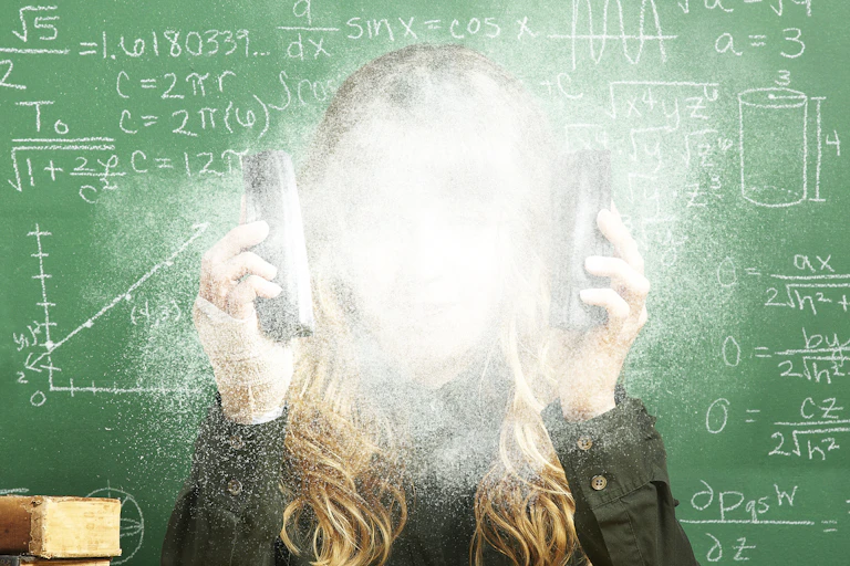 A woman sits at a desk in front of a green chalkboard and holds her hands up to her face, which is covered in chalk dust.