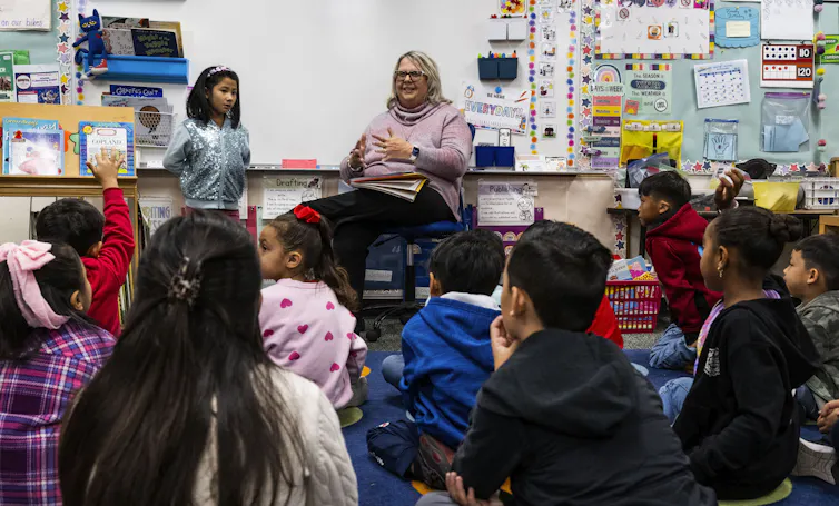 A woman sits in front of a group of young children in a classroom.