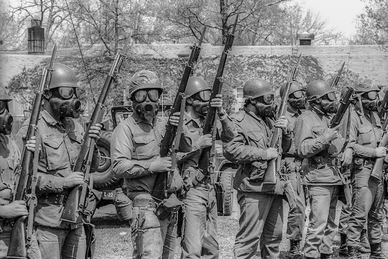 A line of men in military uniforms, wearing gas masks and holding guns.
