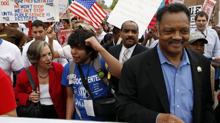 Jesse Jackson smiles bemusedly, holding a sign as he stands in front of other marchers.