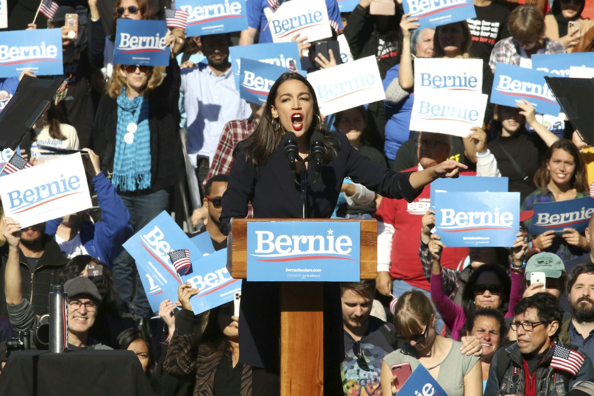 Congresswoman Alexandria Ocasio-Cortez stands at a lectern and appears to shout to a campaign rally crowd.