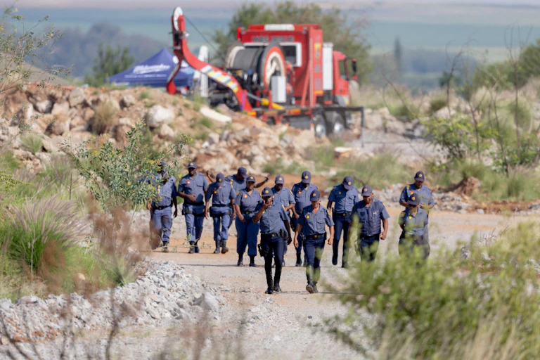 People in blue uniforms walk in a group in a rocky area. In the background is a heavy vehicle with a cable winding mechanism.