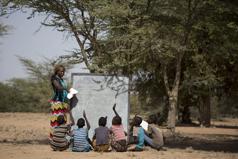 A female teacher instructs children seated under a tree