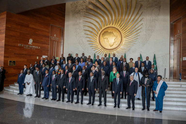 A group of men and women standing on a set of stairs in front of a large gold wall hanging showing a sun with rays and the African map in the middle
