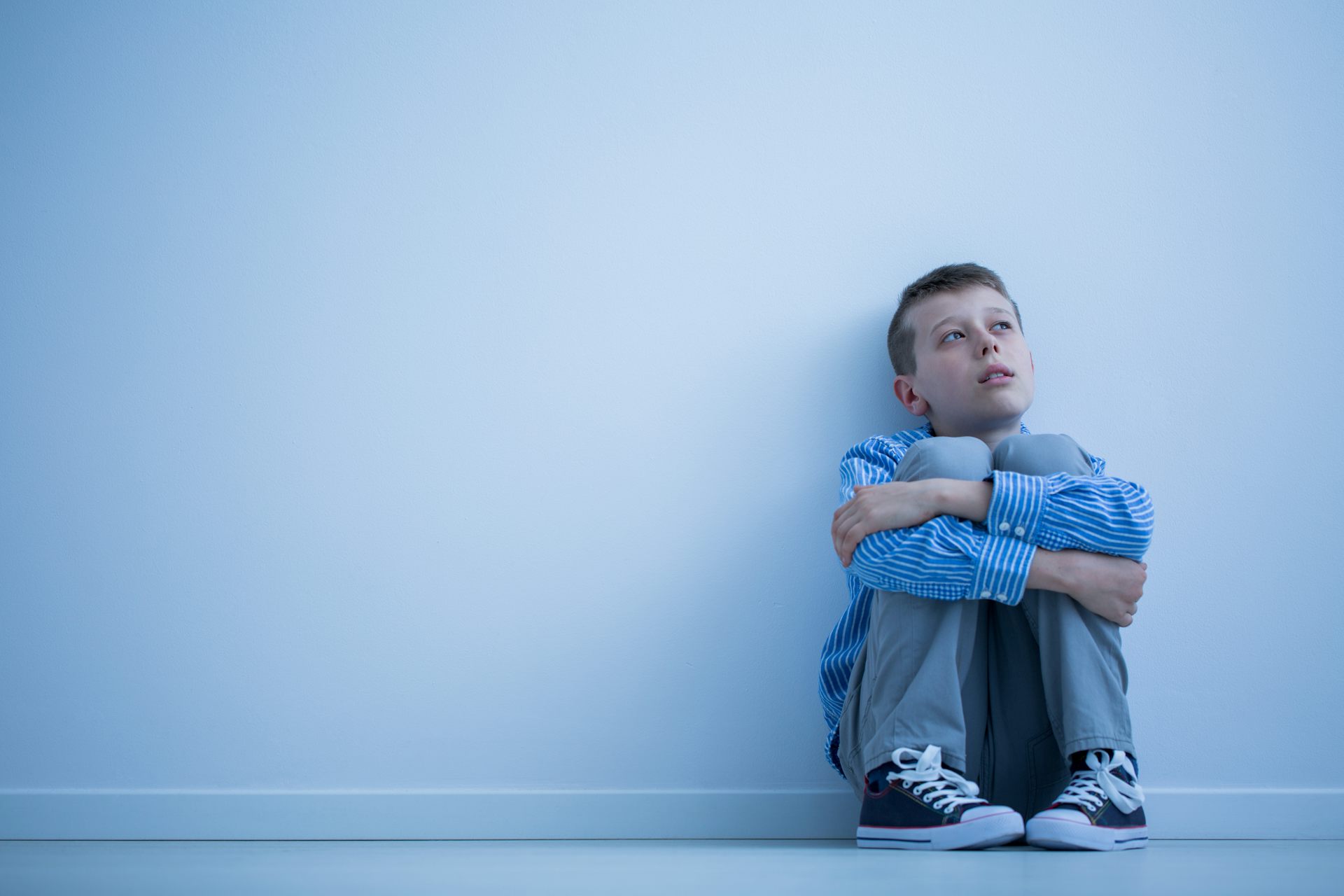 A young boy sits on the floor hugging his knees and looking worried or pensive.