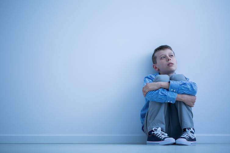 A young boy sits on the floor hugging his knees and looking worried or pensive.