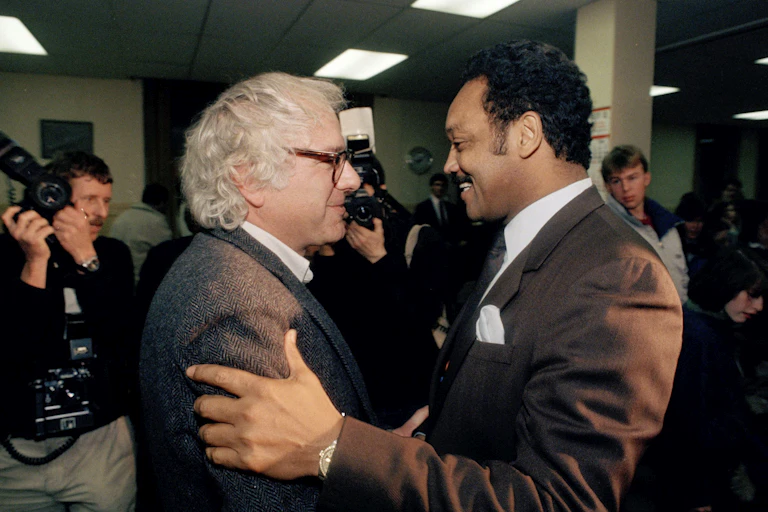 Bernie Sanders and Jesse Jackson stand face to face, Jackson placing his hand on Sanders' right arm.