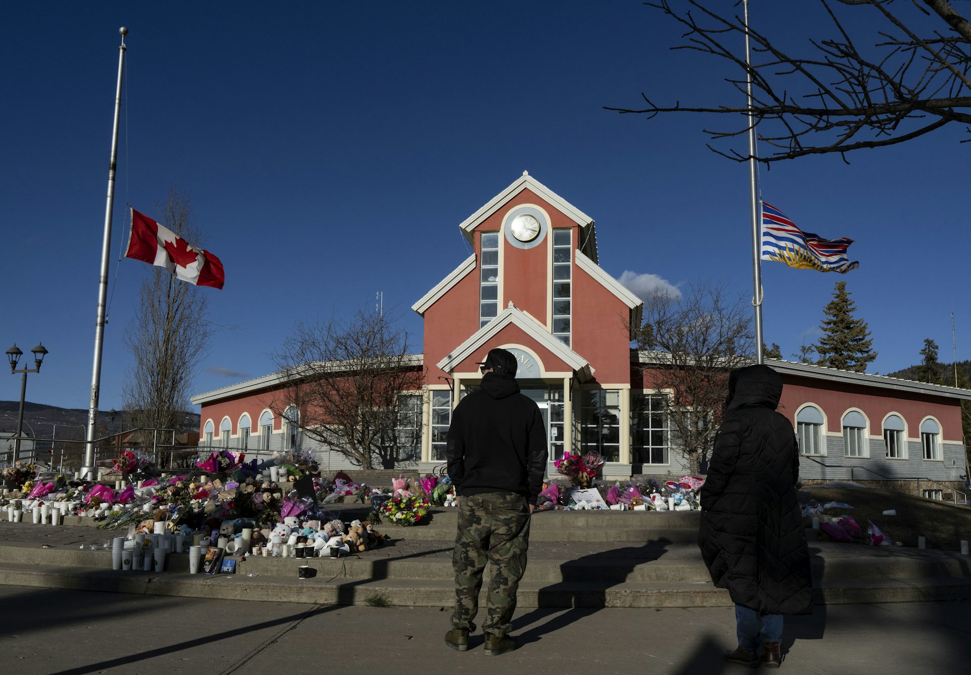 Una persona mira los ramos de flores dejados en un monumento en el suelo, entre dos banderas que ondean a media asta.