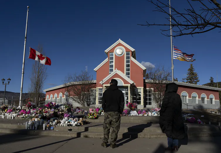 Una persona mira los ramos de flores dejados en un monumento en el suelo, entre dos banderas que ondean a media asta.