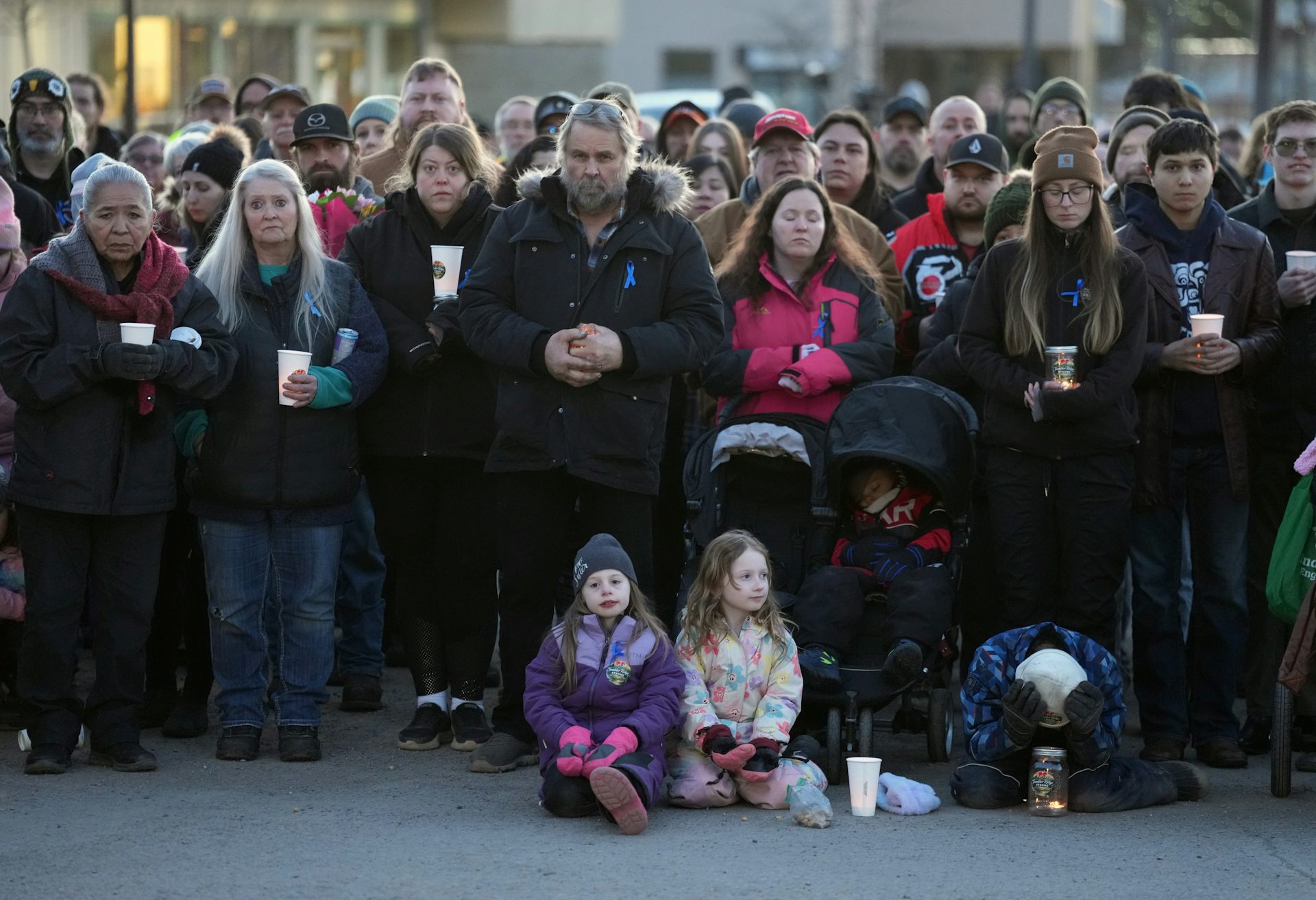 Se ve a un grupo de personas sosteniendo velas, muchas de ellas con los ojos cerrados, y a un niño sentado con las manos sobre la cabeza.