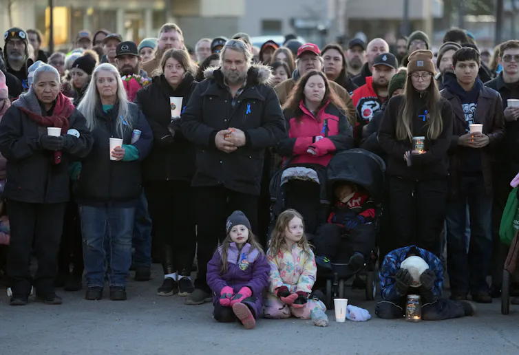 Se ve a un grupo de personas sosteniendo velas, muchas de ellas con los ojos cerrados, y a un niño sentado con las manos sobre la cabeza.