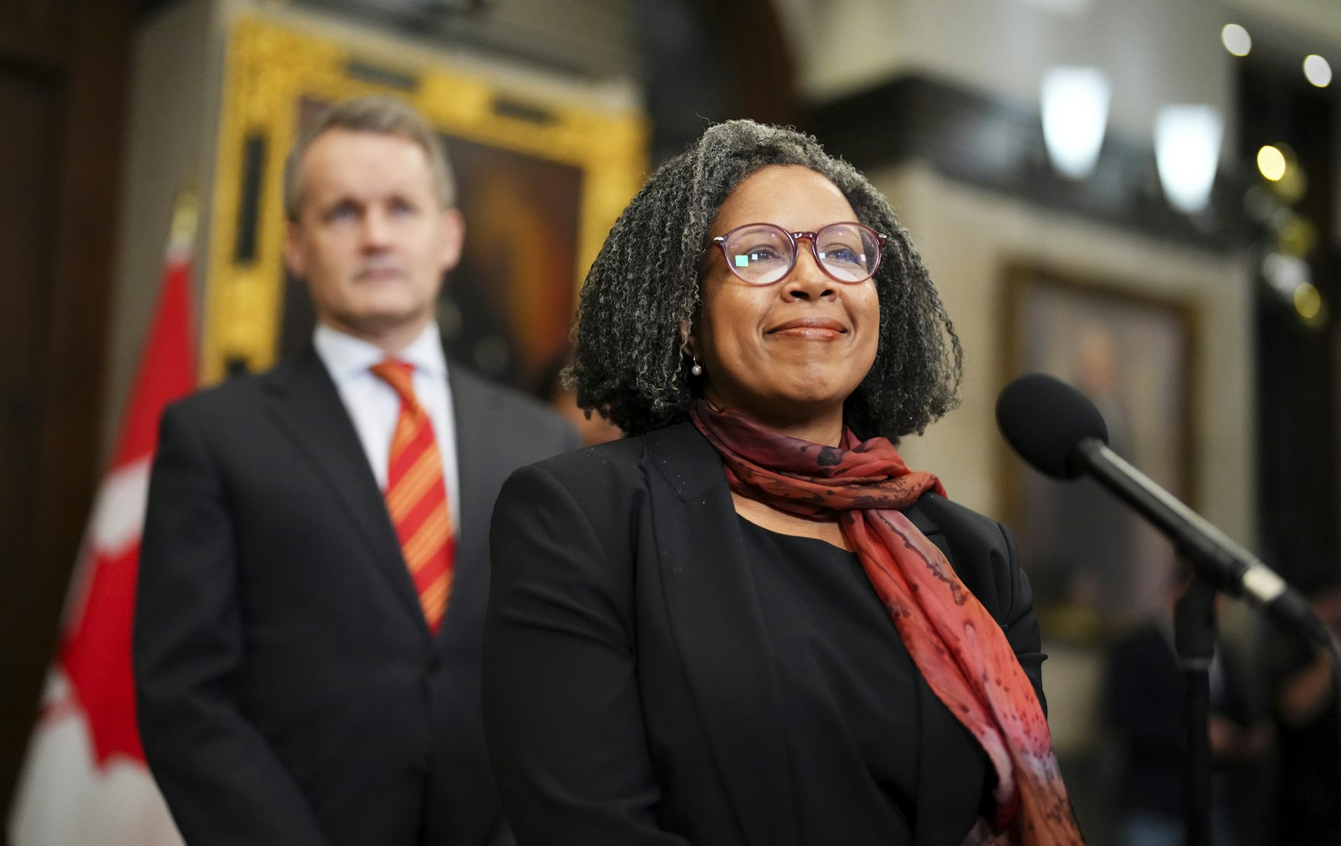 Una mujer negra habla por un micrófono mientras un hombre blanco está detrás de ella.