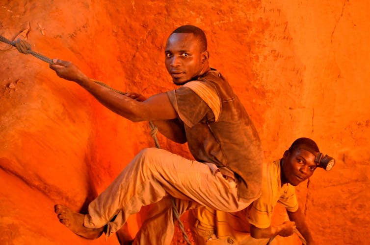 two men climbing out of small mining pit using a rope.