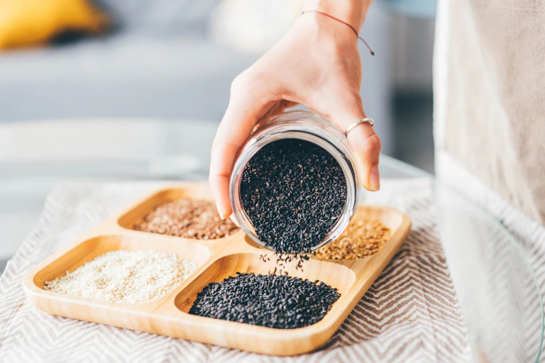 Woman pouring black sesame seeds onto divided plate, each portion containing different seeds, nuts or grains