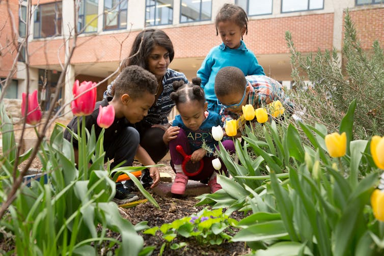 Young children seen in a garden with an adult speaking with them.