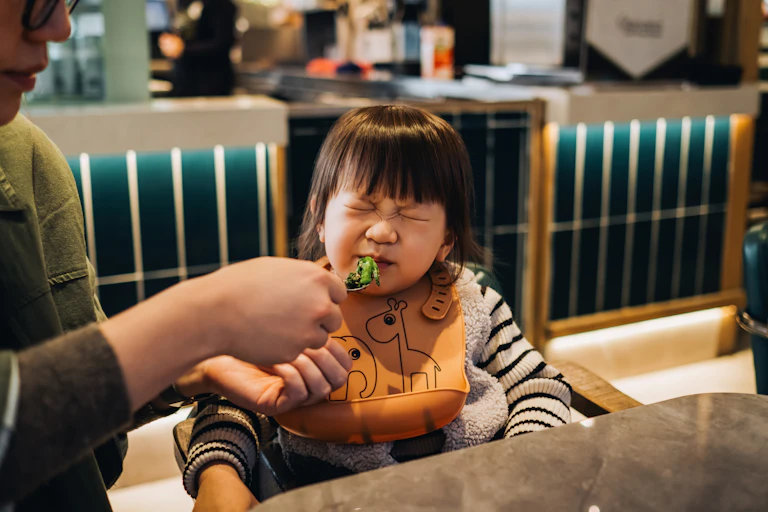 Toddler wearing bib, face scrunched up as parent presents a spoon of greens to them