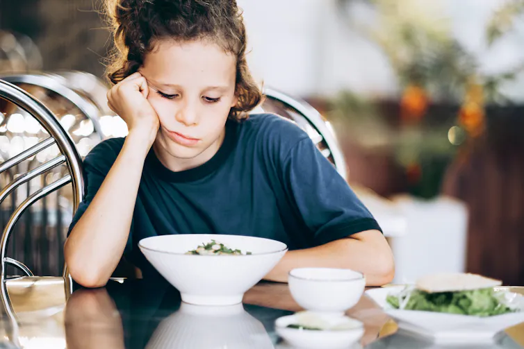 Child looking down at bowl of food with a frown, face propped up against hand on dinner table