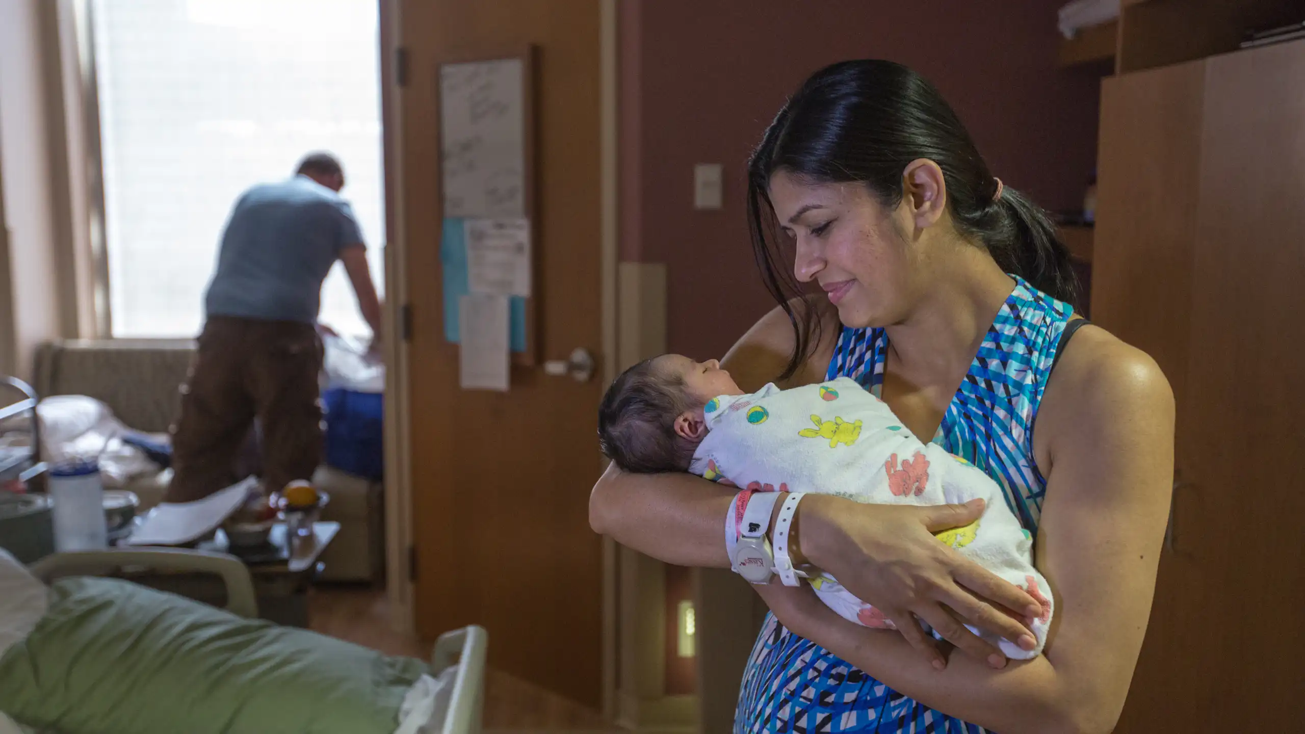 A woman in a blue dress holds a swaddled baby in a hospital room.