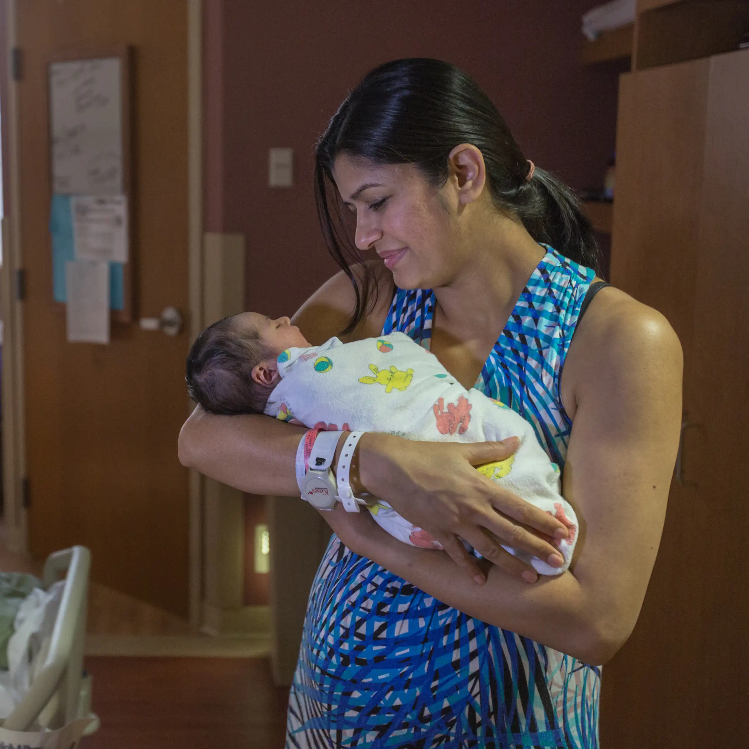 A woman in a blue dress holds a swaddled baby in a hospital room.