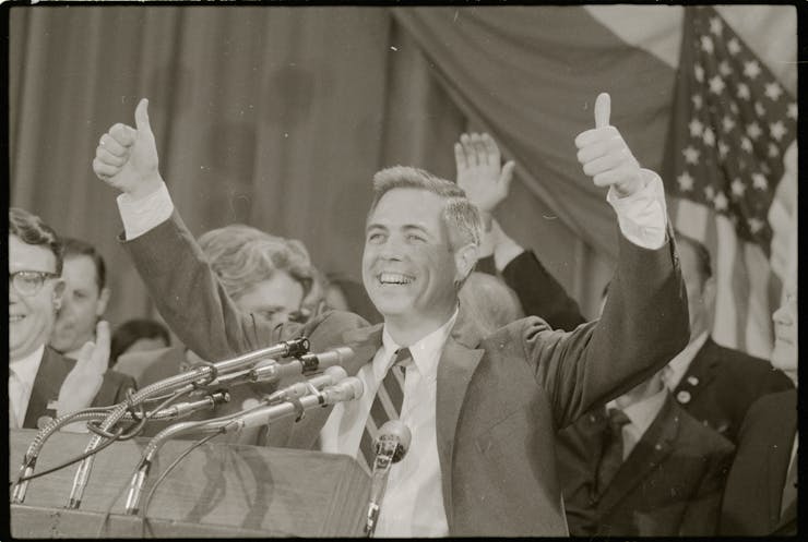 A man in a sports jacket and tie, gives thumbs up as he stands behind a lectern featuring microphones.