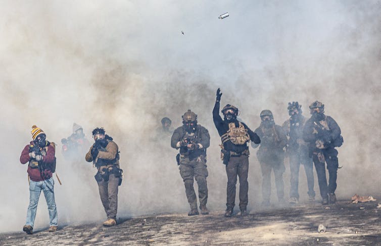 A man dressed in military gear throws a tear gas canister in the air.