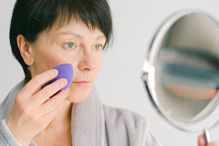 An older woman looks at herself in a mirror while applying makeup