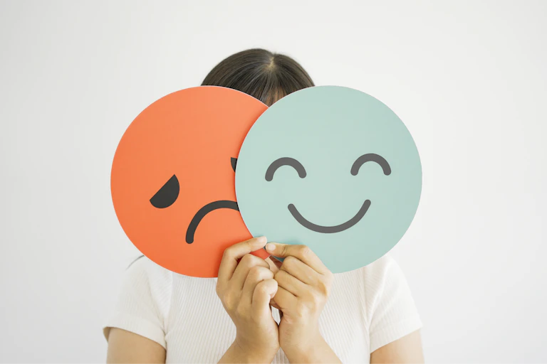A woman holding a cardboard smiley face and a sad face in front of her own face.