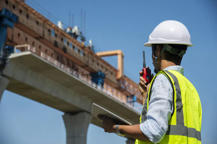 An engineer in a yellow safety vest and white hard hat speaks into a walkie-talkie as he surveys a building site.