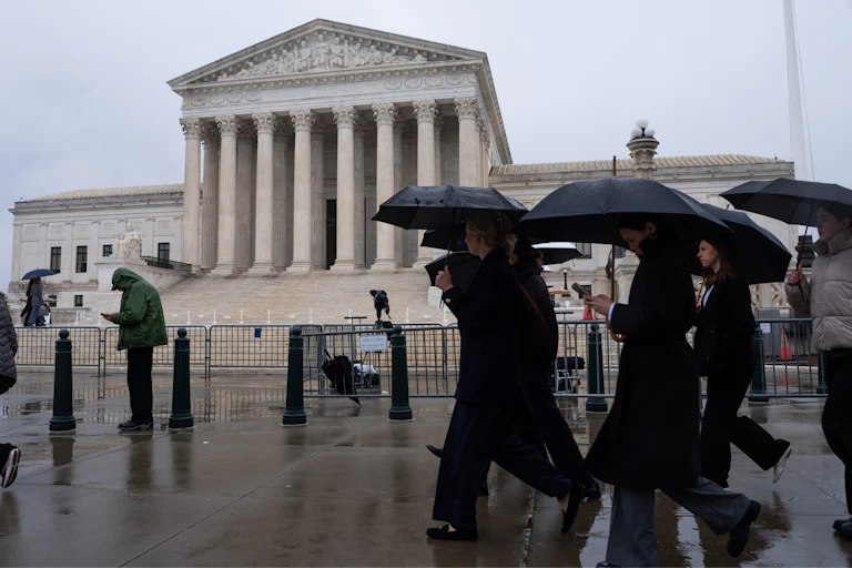 A group of people carry umbrellas as they walk past a big white building.